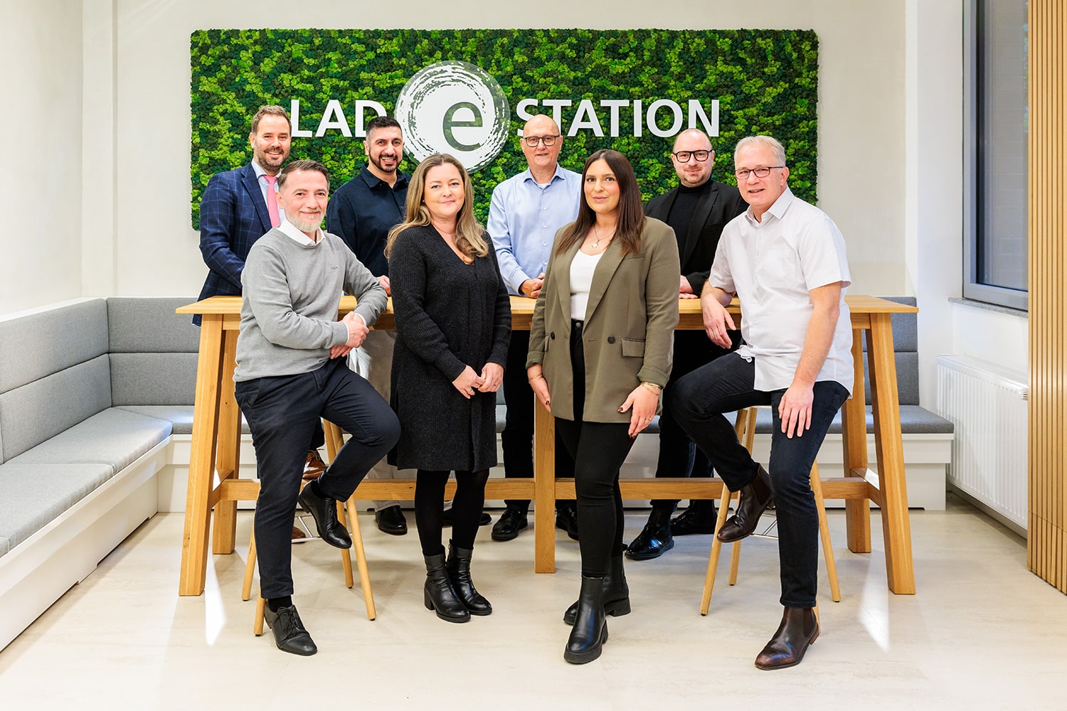 Gruppenfoto des medl Kundenservice-Teams im modernen Pausen- und Besprechungsbereich mit grünem Mooswand-Design und „Ladestation“-Schriftzug.