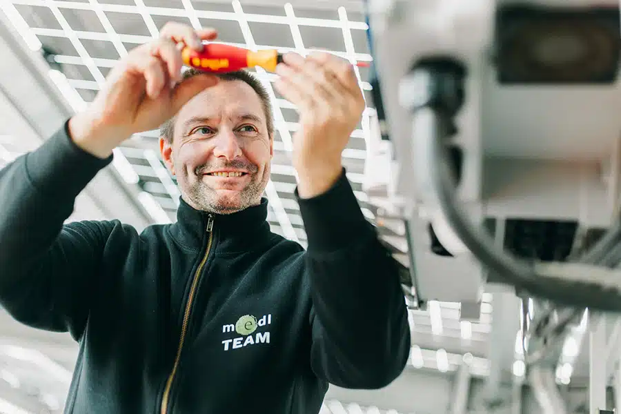 Ein Techniker des medl-Teams bei der Arbeit an einer Photovoltaikanlage, lächelnd und mit Werkzeug in der Hand unter Solarpanelen.